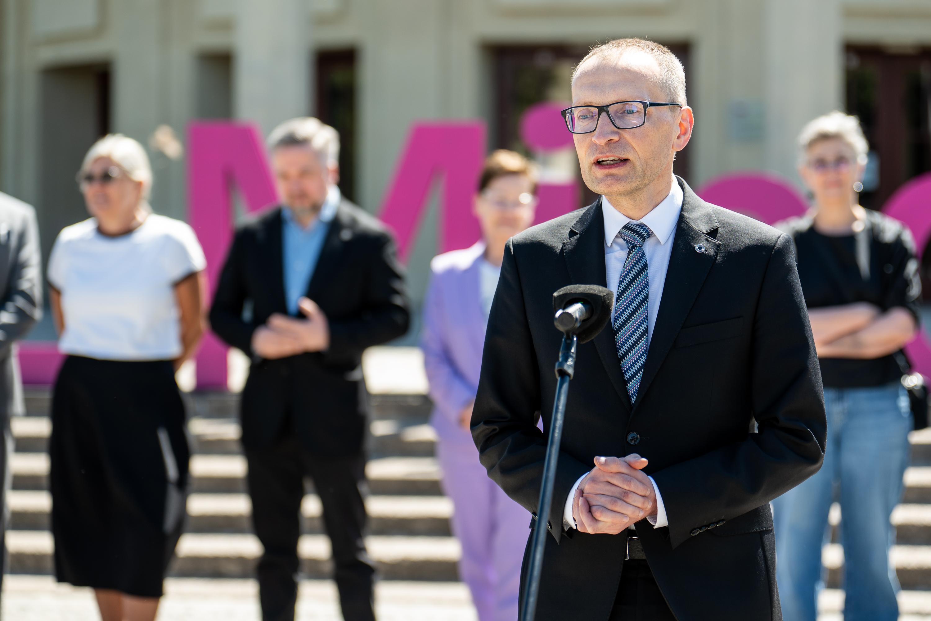 mężczyzna w garniturze przemawia do mikrofonu na placu pod Iglicą przy Hali Stulecia we Wrocławiu, w tle uczestnicy konferencji i duże litery w fioletowo-różowej kolorystyce z napisu Re_Minnd