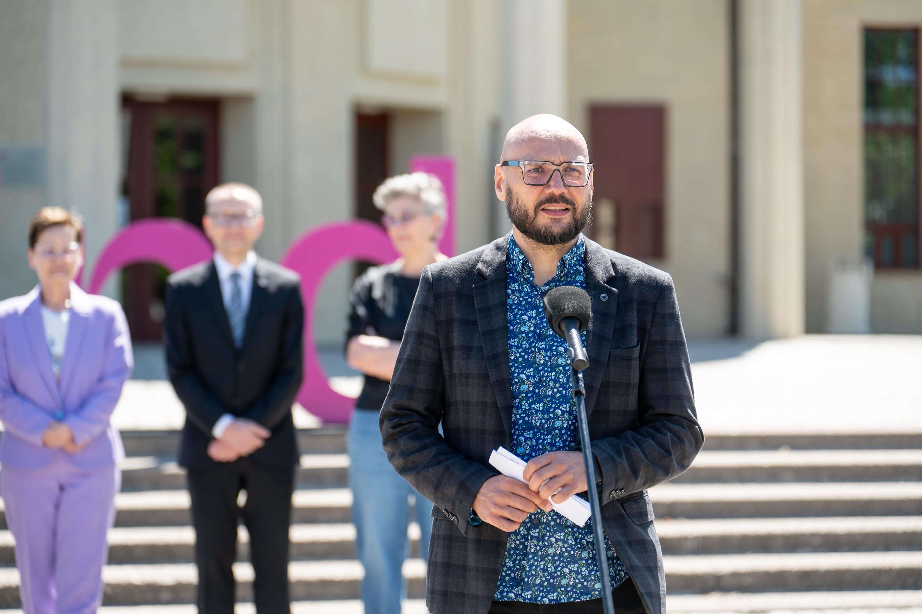 mężczyzna w marynarce i wzorzystej koszuli przemawia do mikrofonu na placu pod Iglicą przy Hali Stulecia we Wrocławiu, w tle uczestnicy konferencji i duże litery w fioletowo-różowej kolorystyce z napisu Re_Minnd