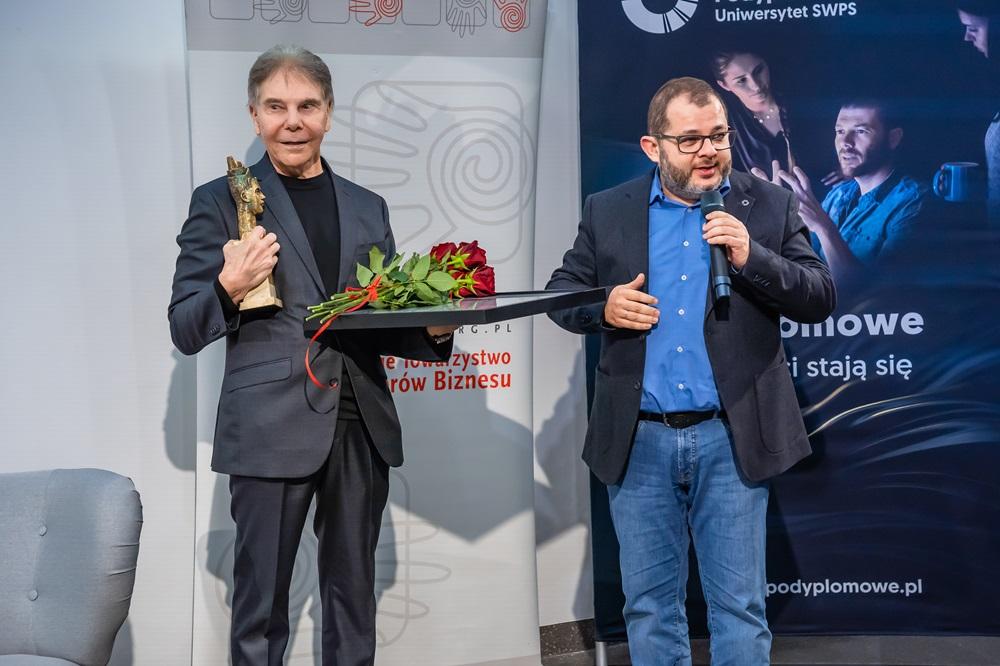 Profesor Cialdini trzymający statuatkę i kwiaty, obok stoi Profesor Tomasz Grzyb z mikrofonem