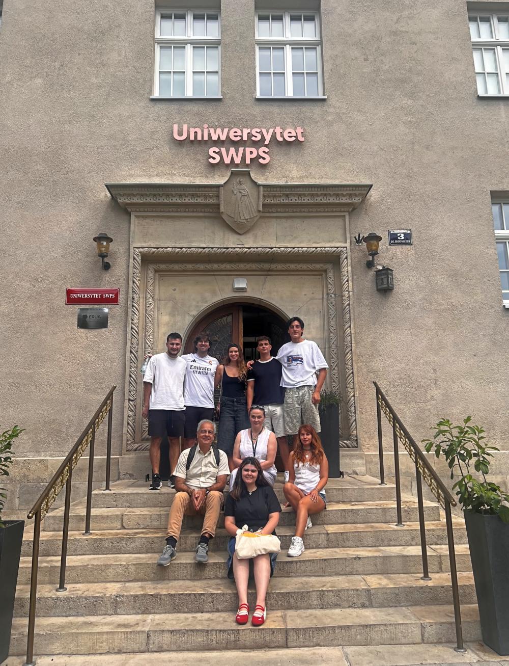 Grupa studentów i wykładowców siedzi na schodach wiodących do budynku USWPS w Krakowie
