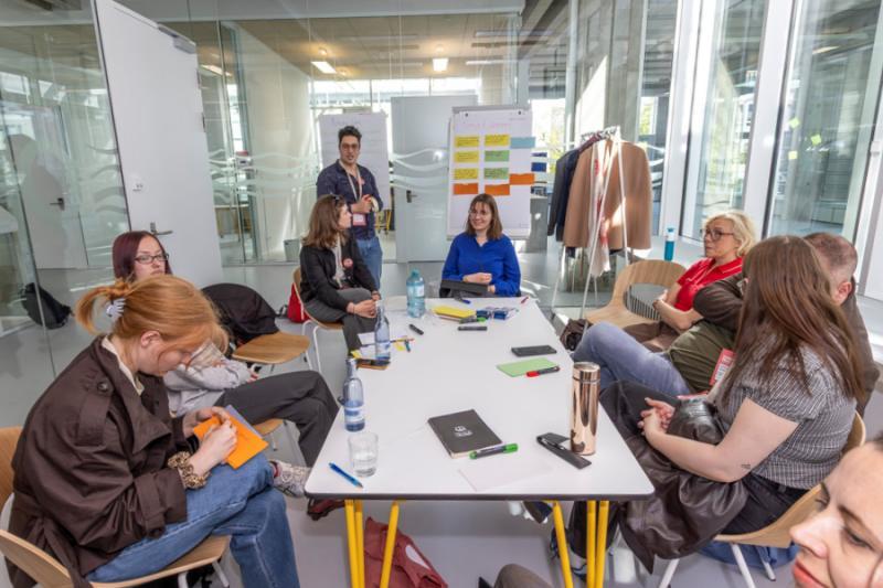 Studenci zgromadzeni wokół stołu słuchają prezentacji i patrzą na flipchart