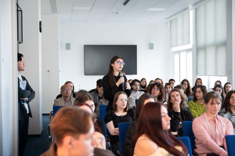 Sala pełna studentów i studentek, jedna ze studentek stoi i mówi przez mikrofon