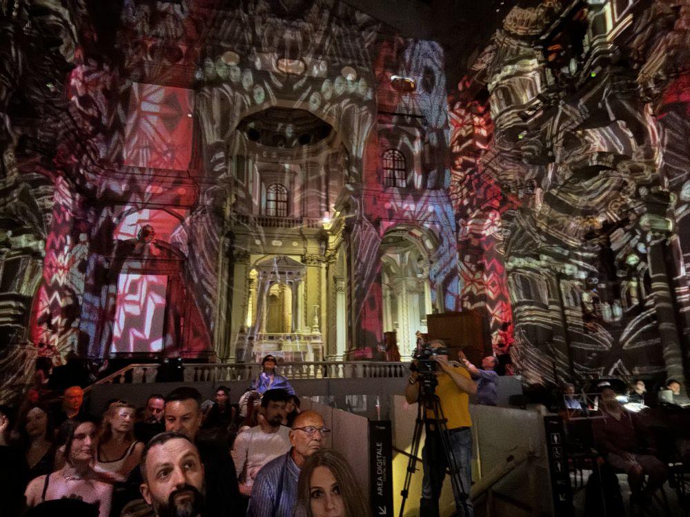 Widownia przedstawienia i kamerzysta we wnętrzu kościoła, na którego ścianach wyświetlane są ozdobne wzory