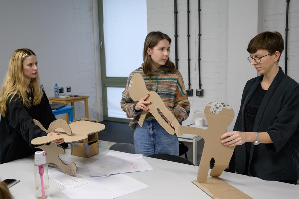 Dwie studentki rozmawiające z prowadzącą warsztaty, wszystkie trzymają w rękach elementy wycięte z kartonu i sklejki. 