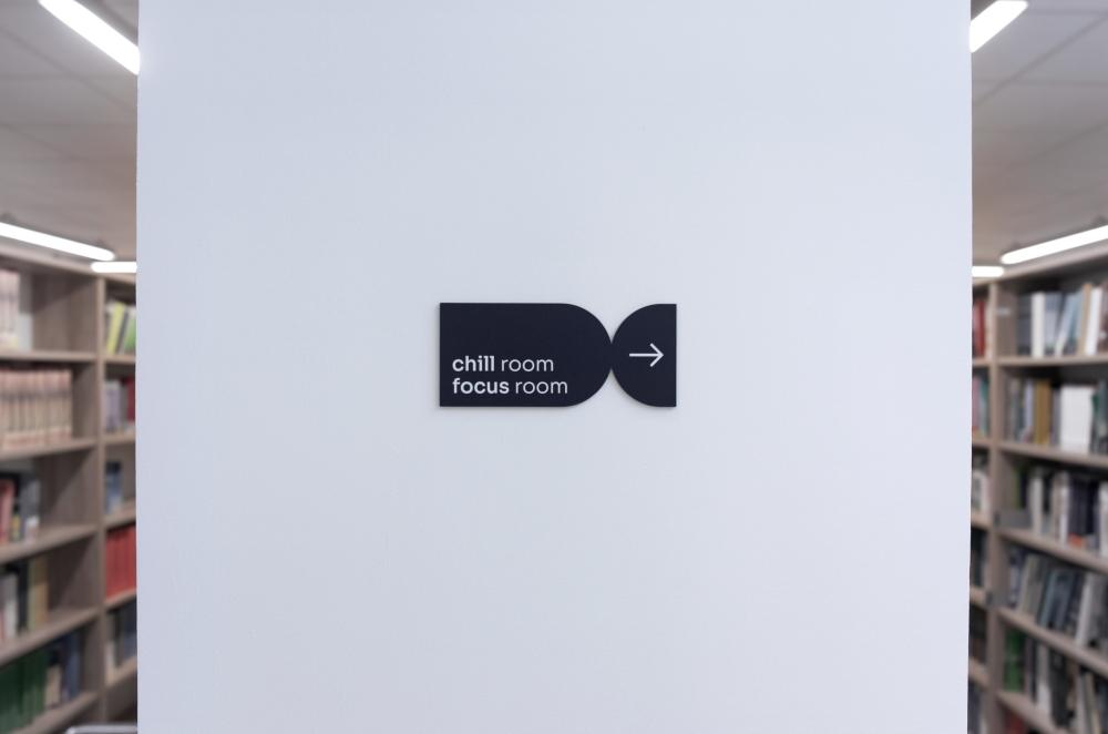 Czarny, poziomy znak kierunkowy na białym filarze z napisem „chill room / focus room” i strzałką w prawo, na tle regałów bibliotecznych.