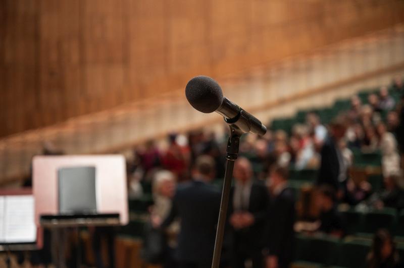 Zbliżenie na mikrofon. W tle widać rozmazane rzędy krzeseł, ludzie zajmują właśnie miejsca na widowni