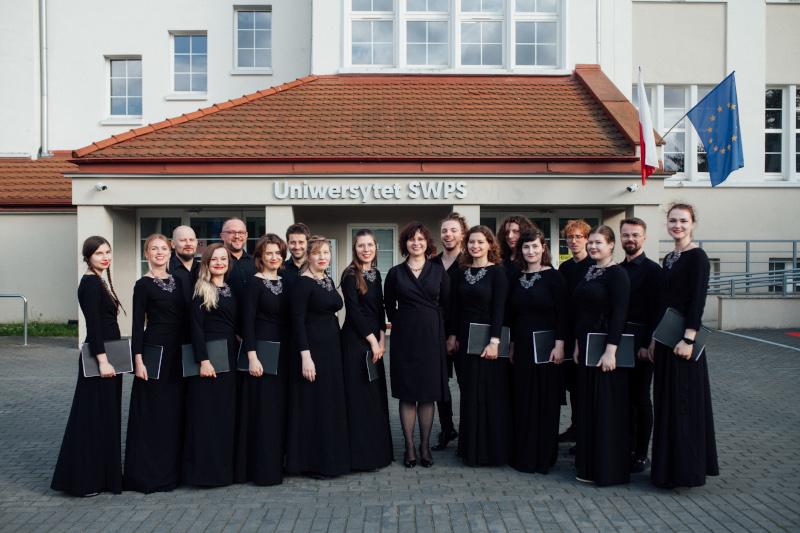 Chórzystki i chórzyści w czarnych strojach chóralnych wraz z dyrygentką, Ewą Mackiewicz, przed siedzibą Uniwersytetu SWPS w Sopocie