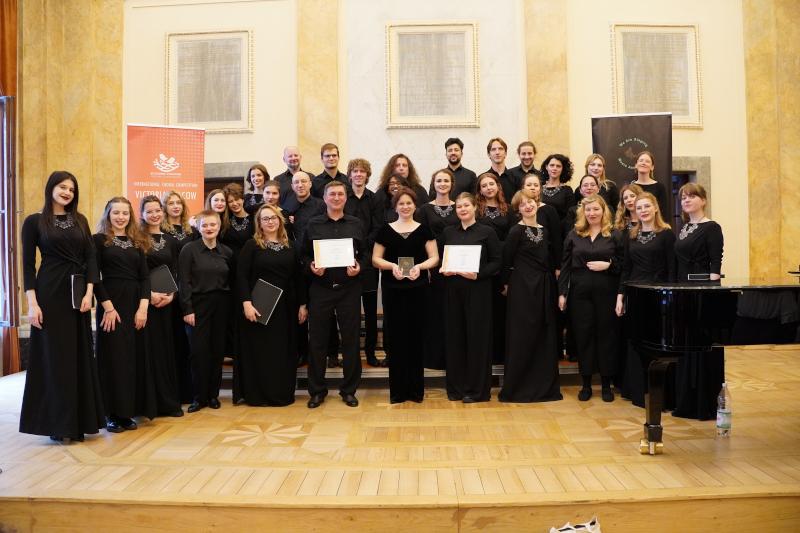 Chór Akademicki Uniwersytetu SWPS na międzynarodowym konkursie Victoria Cracow. Chórzyści i chórzystki stoją  na scenie, w centralnej części zdjęcia, a po prawej stronie jest pianino. Chórzyści mają na sobie czarne sukienki lub czarne koszule i czarne spodnie. Na środku zdjęcia stoi dyrygentka chóru – Ewa Mackiewicz, a po jej prawej i lewej stronie są osoby trzymające dyplomy. Osoby na zdjęciu uśmiechają się