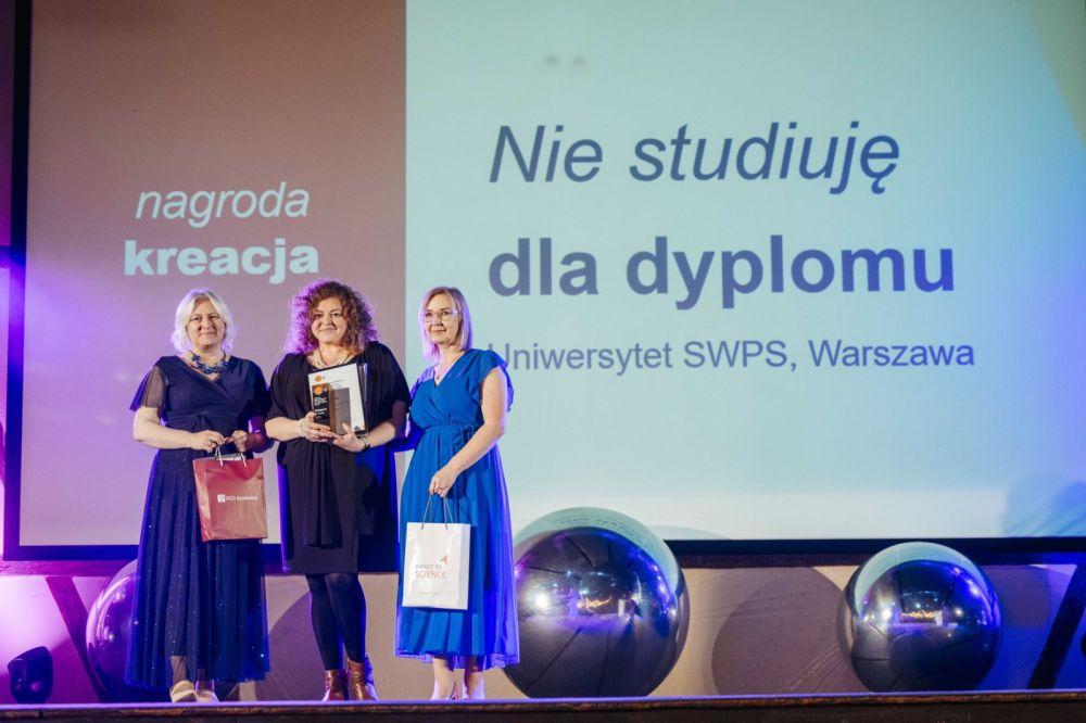 Agnieszka Kampka, Ewelina Kolendowicz-Ossowska i Anna Worosz stoją na scenie. W tle slajd z tekstem „nagroda kreacja” oraz „Nie studiuję dla dyplomu. Uniwersytet SWPS, Warszawa”